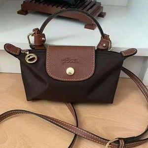 May include: A small, dark brown handbag with a brown leather flap and handle. The bag has a long, adjustable strap and gold-colored hardware. The flap is secured with a gold-colored button and features a logo.