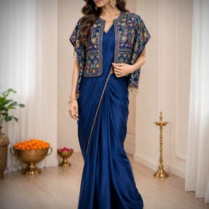 May include: A deep blue satin saree paired with a short, open-front jacket featuring intricate multi-coloured floral embroidery and gold trim. The ensemble includes a matching blue blouse and gold-toned earrings, creating a coordinated look.