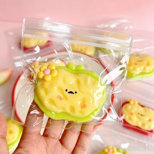 May include: A yellow and green apple-shaped charm with a flower and a cute face, inside a clear plastic bag. The charm has a green border and small orange details. Other similar charms are visible in the background.