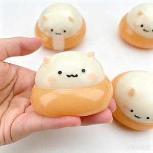 May include: A collection of four cat-shaped toys. Each toy has a white head with small ears and a cartoon face, atop an orange base. The toys are held in a hand.