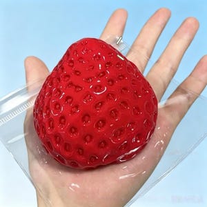May include: A large, bright red strawberry with a textured surface. The strawberry is held in a clear plastic bag, against a light blue backdrop.