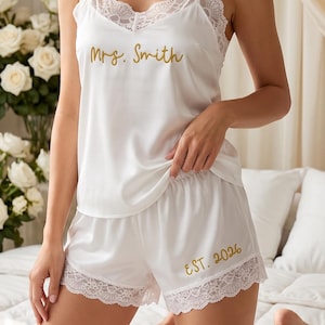 May include: A white satin pyjama set consisting of a camisole and shorts with delicate lace trim. The camisole features the gold embroidered text Mrs. Smith, and the shorts feature the gold embroidered text EST. 2026 on the left leg.