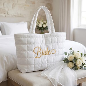 May include: A white quilted tote bag featuring the word Bride embroidered in gold script lettering on the front. The bag has two padded handles and a rectangular structure, accompanied by a bouquet of white roses and a sheer white veil.