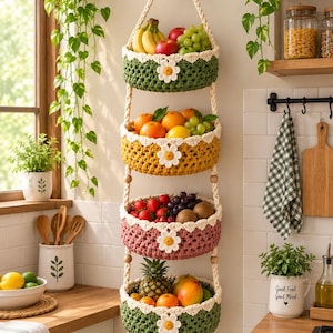 Puede incluir: Una cesta de frutas colgante de cuatro niveles hecha de hilo de ganchillo en verde, amarillo y rosa, con margaritas blancas en cada cesta. Los niveles están unidos por una cuerda gruesa color crema con cuentas de madera y dos borlas.