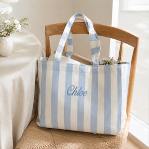 May include: A rectangular tote bag featuring vertical light blue and white stripes. The bag has two matching striped shoulder straps and the name Chloe embroidered in blue script lettering centered on the front panel of the fabric exterior.