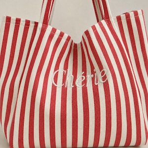May include: A tote bag featuring a vertical red and white striped pattern. The word "Ch&eacute;rie" is embroidered in white cursive script across the centre of the bag. It includes two matching red and white striped fabric handles at the top.
