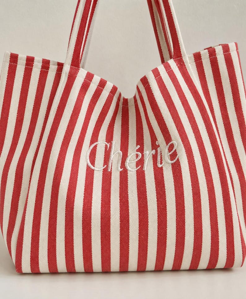 May include: A tote bag featuring a vertical red and white striped pattern. The word "Ch&eacute;rie" is embroidered in white cursive script across the centre of the bag. It includes two matching red and white striped fabric handles at the top.