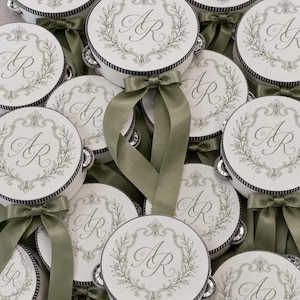 May include: A collection of round white tambourines featuring a black striped border and a central olive green laurel wreath design surrounding the monogram letters AR. Each tambourine is adorned with a tied olive green satin ribbon bow.