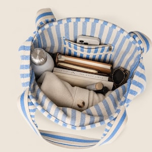 May include: A tote bag featuring a blue and white vertical striped pattern. The interior is filled with various items including a silver water bottle, a smartphone in a pocket, two notebooks, a pair of sunglasses, and a folded beige garment.
