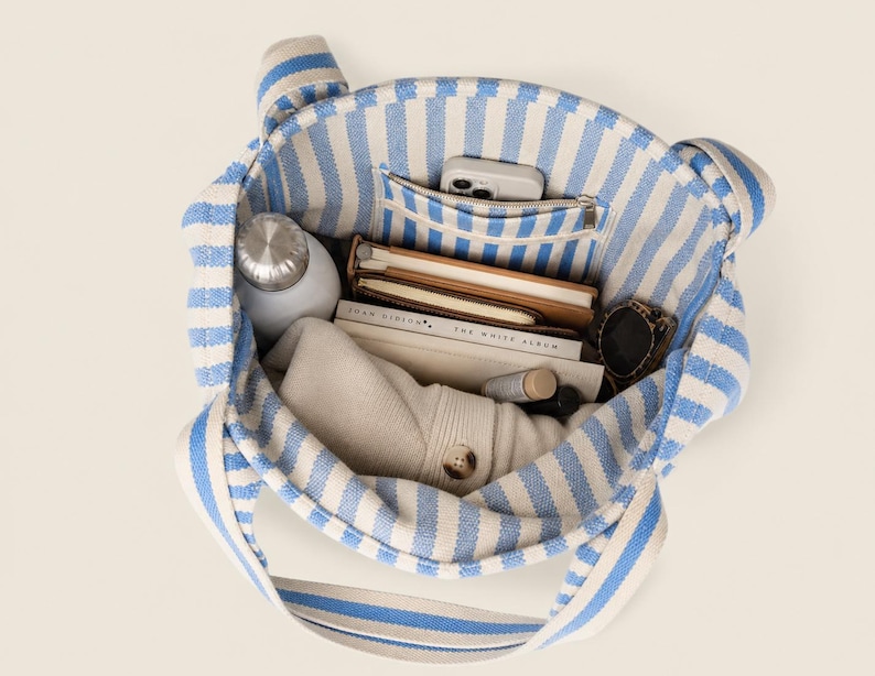May include: A tote bag featuring a blue and white vertical striped pattern. The interior is filled with various items including a silver water bottle, a smartphone in a pocket, two notebooks, a pair of sunglasses, and a folded beige garment.