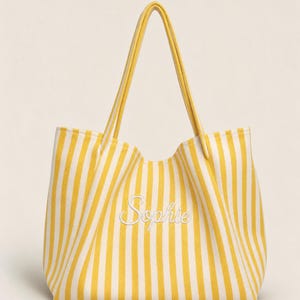 May include: A yellow and white vertical striped tote bag featuring two long shoulder straps and the name Sophie embroidered in white cursive script across the centre of the front panel. The bag has a structured base and open top design.