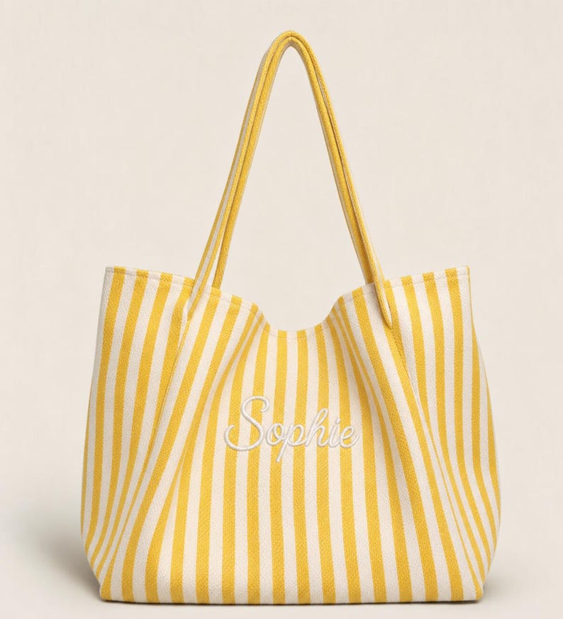 May include: A yellow and white vertical striped tote bag featuring two long shoulder straps and the name Sophie embroidered in white cursive script across the centre of the front panel. The bag has a structured base and open top design.