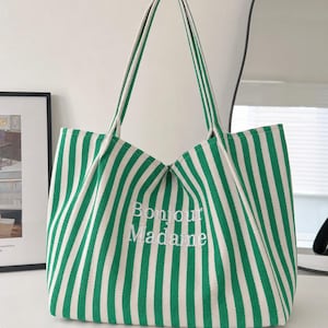 May include: A green and white vertically striped canvas tote bag featuring two shoulder straps and the embroidered white text Bonjour Madame centred on the front. The bag has a structured base and an open top design with a fabric construction.