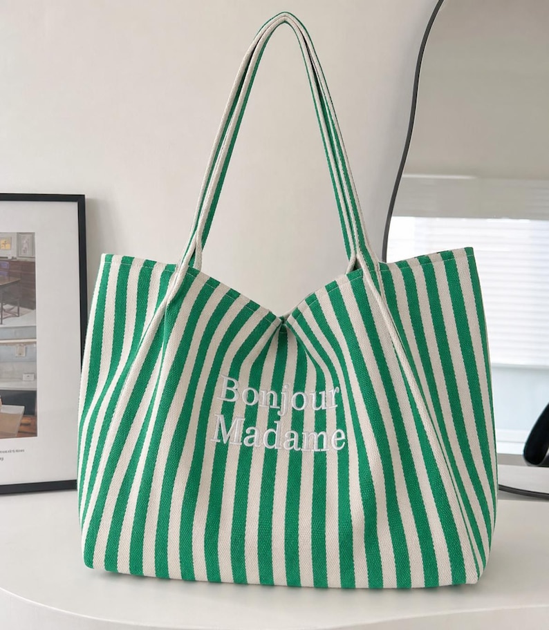 May include: A green and white vertically striped canvas tote bag featuring two shoulder straps and the embroidered white text Bonjour Madame centred on the front. The bag has a structured base and an open top design with a fabric construction.