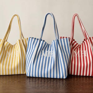 May include: Three tote bags with vertical stripes in yellow, blue, and red, each featuring two shoulder straps and the white embroidered monogram letters MH centred on the front of the fabric body of every individual bag.