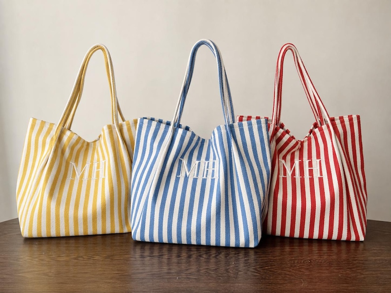 May include: Three tote bags with vertical stripes in yellow, blue, and red, each featuring two shoulder straps and the white embroidered monogram letters MH centred on the front of the fabric body of every individual bag.