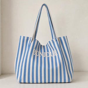 May include: A tote bag featuring a vertical blue and white striped pattern. The word BONJOUR is embroidered in white capital letters across the centre of the bag. It has two long shoulder straps attached to the top edge of the rectangular body.