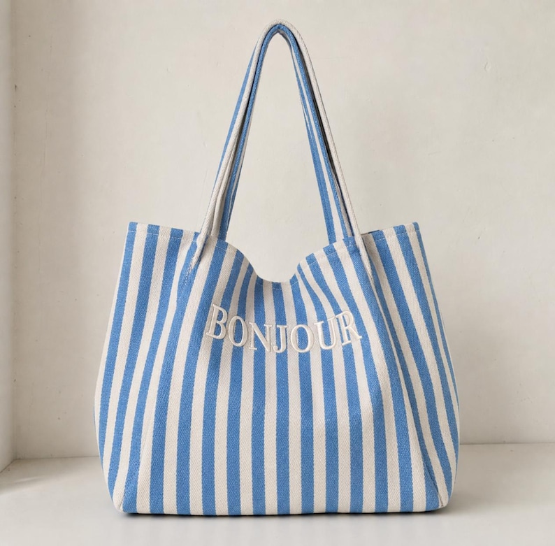 May include: A tote bag featuring a vertical blue and white striped pattern. The word BONJOUR is embroidered in white capital letters across the centre of the bag. It has two long shoulder straps attached to the top edge of the rectangular body.