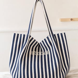 May include: A navy blue and white striped canvas tote bag with two long shoulder straps. The word H&Eacute;RITAGE is embroidered in white thread on the front centre of the bag. The bag features a structured base and a vertical striped pattern throughout.