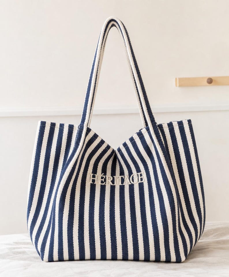 May include: A navy blue and white striped canvas tote bag with two long shoulder straps. The word H&Eacute;RITAGE is embroidered in white thread on the front centre of the bag. The bag features a structured base and a vertical striped pattern throughout.