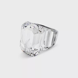 Op de afbeelding: Een grote, heldere, rechthoekig geslepen ring met een gefacetteerd oppervlak. De ring is gemaakt van een transparant materiaal, mogelijk kristal of glas, en ligt op een witte ondergrond. Het ontwerp van de ring heeft een prominente, geometrische vorm.