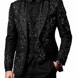 Op de afbeelding: Een zwart driedelig pak bestaande uit een blazer en gilet, versierd met ingewikkeld zwart kraalwerk en borduursels in een bloemmotief. De set bevat een bijpassende zwarte vlinderdas, een zwart overhemd en een zwarte pantalon.