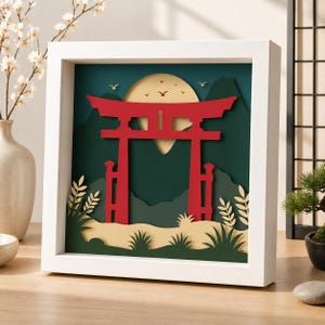 May include: A square white shadow box frame containing a layered paper art piece. The scene features a bright red torii gate centered in front of a large pale yellow moon, dark green mountain silhouettes, and stylized green foliage in the foreground.