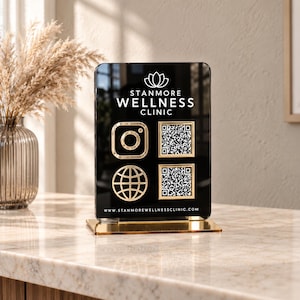 May include: A rectangular black acrylic sign with a gold base featuring the white text STANMORE WELLNESS CLINIC, a lotus logo, an Instagram icon, a globe icon, two QR codes, and the website address www.stanmorewellnessclinic.com printed in white.