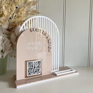 Peut inclure: Un panneau en acrylique beige et blanc avec un cadre arqué blanc, le texte "LEAVE US A REVIEW" et "artistik beauty the brow specialist", ainsi qu'un QR code carré à bordure or rose, fixé sur une base rectangulaire avec un petit plateau blanc.
