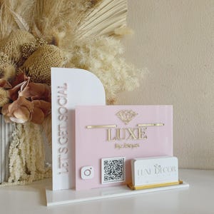 Op de afbeelding: Een op maat gemaakt acrylbord met een wit boogvormig paneel met de tekst LET'S GET SOCIAL en een roze rechthoekig paneel met een diamanten icoon, de tekst LUXE BY JESSY, een Instagram-logo, een QR-code en een visitekaarthouder met LUXE DECOR.