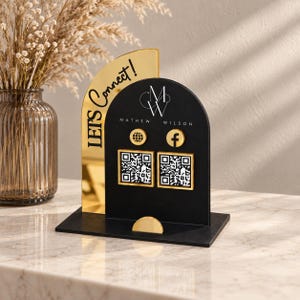 May include: A black and gold acrylic social media sign featuring a semi-circular black panel with the text Mathew Wilson, a logo, two circular icons for a website and Facebook, and two square QR codes. A gold arched side panel reads Let's Connect!