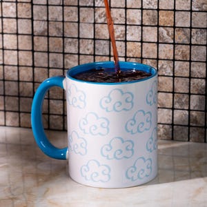 Può includere: Una tazza in ceramica bianca con manico e interno blu brillante, caratterizzata da un motivo ripetuto di nuvole azzurre sull'esterno. Un liquido marrone scuro viene versato nella tazza, riempiendola fino all'orlo con una bevanda scura e ricca.