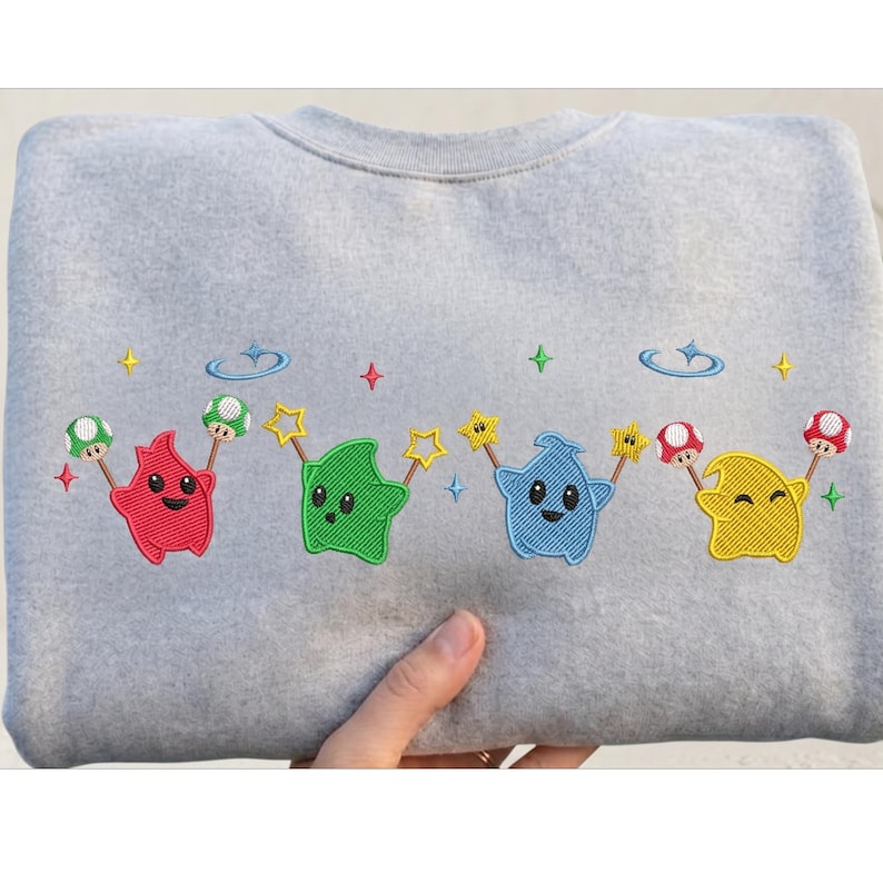 Puede incluir: Sudadera gris claro con personajes de dibujos animados bordados en rojo, verde, azul y amarillo. Cada personaje sostiene una varita de estrella y una seta. El dise&ntilde;o incluye estrellas y nubes.