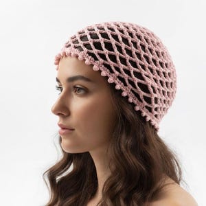 Pode incluir: Um gorro de crochê rosa claro com um padrão de malha aberta em forma de diamante em toda a coroa. A borda inferior do gorro é finalizada com uma fileira delicada de pequenos pontos redondos e texturizados, espaçados uniformemente.