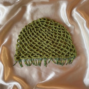 Peut inclure: Un bonnet en crochet vert olive présentant un motif de mailles ajourées en forme de filet sur tout le corps. Le bord inférieur est agrémenté d'une frange de petites perles de rocaille vertes et vert clair suspendues aux boucles.