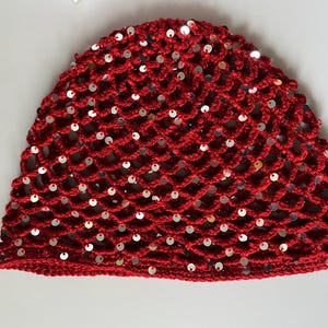 Peut inclure: Un bonnet en crochet rouge présentant un motif en maille ajourée en forme de losanges. Le design est orné de nombreuses petites paillettes argentées, rondes et réfléchissantes, fixées au fil à chaque intersection de la grille.