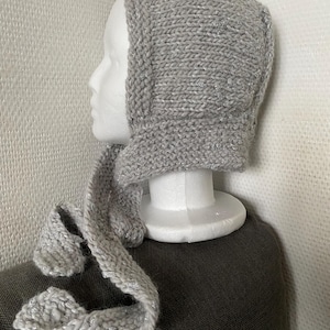 Puede incluir: Un gorro tejido de color gris claro con un sutil hilo metálico plateado entretejido en la lana. El gorro presenta un patrón de punto grueso y texturizado, e incluye dos lazos tejidos largos a juego que cuelgan de los lados.