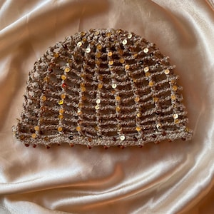 Può includere: Un berretto all'uncinetto color beige con un motivo a rete a maglia aperta, impreziosito da piccole paillettes dorate riflettenti. Il bordo inferiore del cappello è rifinito con una fila di piccole perline marrone scuro lungo l'orlo.