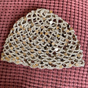Op de afbeelding: Een beige gehaakte muts met een open breipatroon, versierd met talloze kleine, ronde, reflecterende goudkleurige lovertjes die over het gehele oppervlak van het hoofddeksel zijn verspreid, wat zorgt voor een glinsterende en textuurrijke uitstraling.