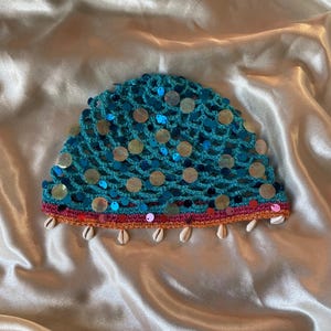 May include: A turquoise crochet beanie featuring a net-like pattern adorned with iridescent circular sequins. The bottom edge is finished with a pink and orange band, and the hem is decorated with a row of small white cowrie shells.