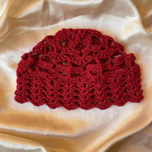 Peut inclure: Un bonnet au crochet rouge vif présentant un motif de points ajourés complexe avec un bord festonné. Le fil présente un léger éclat métallique, créant une apparence texturée et légère avec une calotte arrondie et un design décoratif.