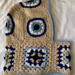 May include: A crochet balaclava constructed from square motifs featuring a central eye pattern. The design incorporates beige, white, dark blue, and light blue yarn colours. The edges are finished with a light blue trim around the face opening.