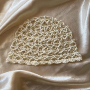 Puede incluir: Un gorro de ganchillo de color crema que presenta un patrón de celosía calado en todo el cuerpo. El gorro está construido con un borde festoneado a lo largo del dobladillo inferior e incorpora pequeñas cuentas transparentes tejidas.