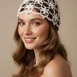 May include: A white crochet cap featuring a delicate floral pattern throughout the openwork design. The headpiece is embellished with small, round white pearls integrated into the centre of each flower and along the scalloped bottom edge.