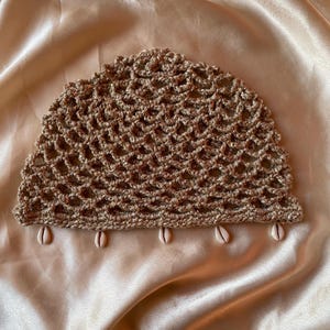 May include: A semi-circular crochet headpiece in a metallic gold tone featuring an open-work mesh pattern. The bottom edge is finished with a straight border adorned with five small, white, oval-shaped cowrie shells spaced evenly along the base.