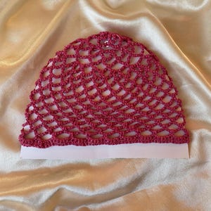 Pode incluir: Um gorro de crochê magenta vibrante com um padrão de pontos abertos tipo rede. O fio apresenta um brilho metálico subtil em toda a sua construção. O gorro é exibido estendido sobre uma superfície de tecido liso e de cor clara.