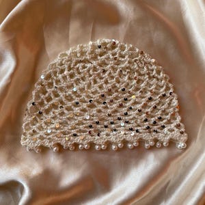 Può includere: Un berretto all'uncinetto color crema caratterizzato da un motivo a rombi a maglia aperta, impreziosito da piccole paillettes argentate riflettenti su tutto il tessuto. Il bordo inferiore del cappello è rifinito con perline bianche pendenti.