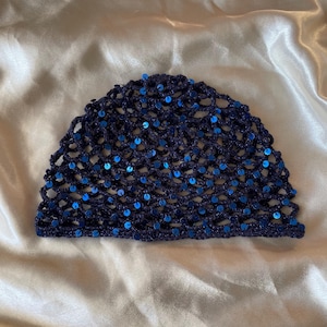Peut inclure: Un bonnet en crochet bleu marine foncé présentant un motif en maille ajourée orné de petites paillettes bleues circulaires et réfléchissantes sur toute la structure. Le bonnet possède un sommet arrondi et un bord inférieur droit.