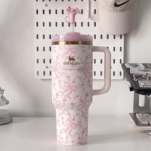 Könnte beinhalten: Ein weißer Stanley LoveShackFancy-Becher mit einem rosa Blumen- und Schleifenmuster, einem goldfarbenen Rand, einem hellrosa Griff und einem passenden rosa Deckel mit einem Strohhalm, der eine kleine rosa Schleife als Aufsatz hat.