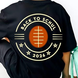 May include: A black t-shirt featuring a graphic on the back with the text BACK TO SCHUL 2026, a brown football illustration, and star accents. The shirt is paired with a black baseball cap, a light green backpack, and a light green skateboard.